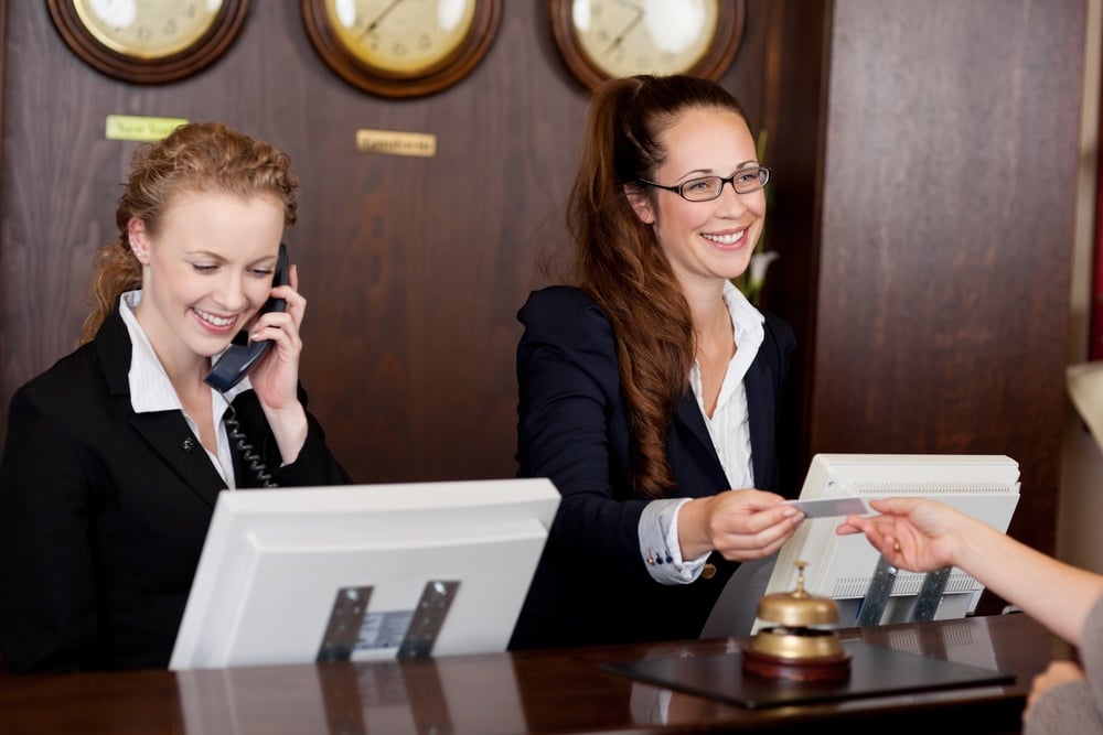 Two beautiful young stylish receptionists at a reception desk, one talking on the telephone and the other handing a card to a customer
